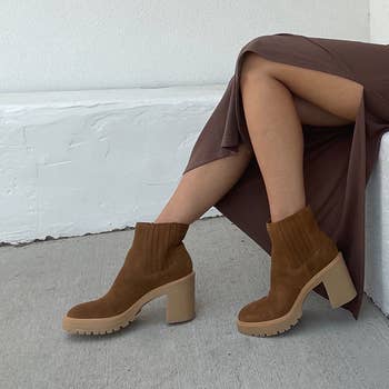 Woman's legs successful  a slit skirt, wearing brownish  ankle boots with chunky heels and ribbed details, sitting connected  a bench.