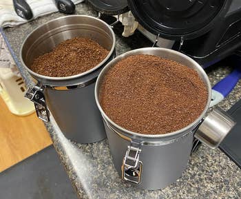 Two coffee canisters with fresh grounds, lids open, on a kitchen counter