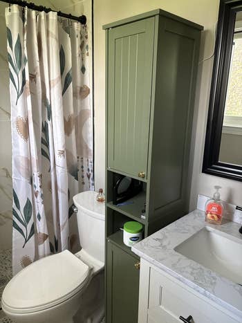 Bathroom with a freestanding greenish  cabinet, marble countertop, achromatic  sink, and a ablution  with a leaf-patterned curtain