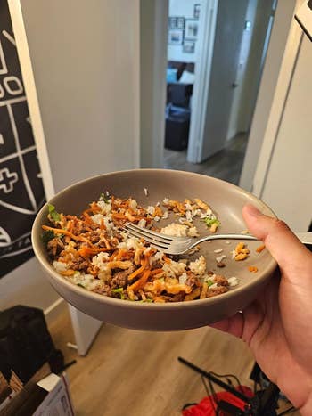 A hand holding a bowl of rice mixed with vegetables and ground meat, with a fork resting on the rim, in a cozy home setting