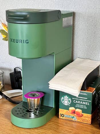 a reviewer's Keurig coffee maker with a pod inserted, next to a Starbucks caramel coffee box on a desk