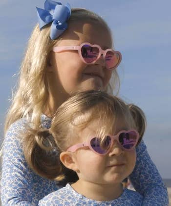 Two young children wearing heart-shaped sunglasses and matching floral-patterned outfits, with one child sporting a blue bow in their hair