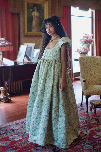 Model wearing a floral-patterned, floor-length dress stands in an ornate room with vintage decor, including framed portraits, a rug, and a piano