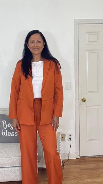 reviewer in orange suit with white top and boots stands in room, near couch with decorative pillow