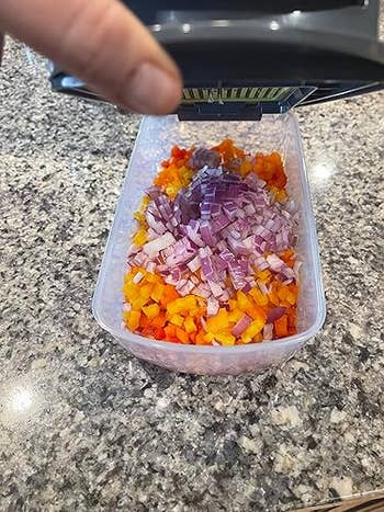 Chopped red onions and peppers are being placed into a clear container with a food chopper on a kitchen countertop