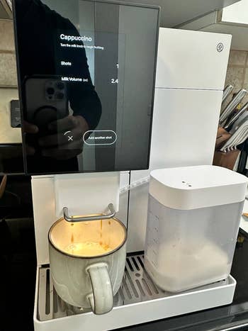 Automatic espresso machine brewing cappuccino, displaying settings on a touchscreen interface. A ceramic mug is placed under the dispenser