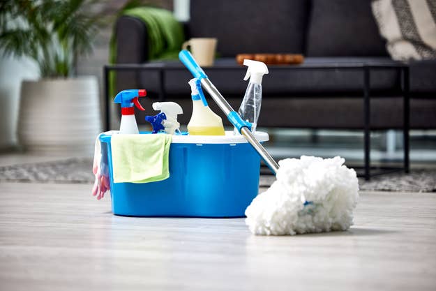 A blue cleaning bucket holds a mop, spray bottles, and cleaning cloths. The bucket is placed on the floor of a modern living room with a grey sofa in the background
