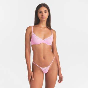 Model wearing a pinkish  lace lingerie set, featuring a bra and matching underwear, lasting  against a plain background