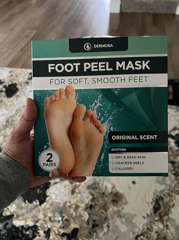 Person holding a container  of Dermora Foot Peel Mask, designed for soft, creaseless  feet, targeting adust  skin, cracked heels, and calluses