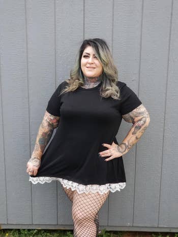 reviewer in a black T-shirt dress with lace hem, fishnet tights, and tattoos, posing with hands on hips