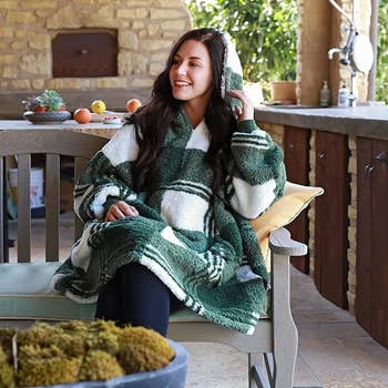 Model sitting on an outdoor bench while wearing the green and white plaid blanket hoodie