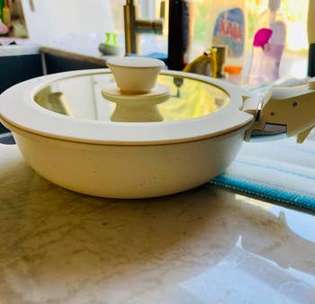 A kitchen scene featuring a ceramic skillet with a glass lid on a countertop, surrounded by dish soap and other cleaning products