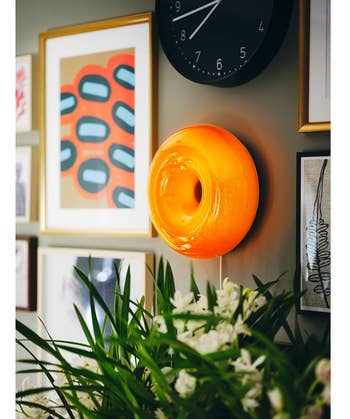 Wall with eclectic framed art, an orangish circular airy fixture, and greenish plants
