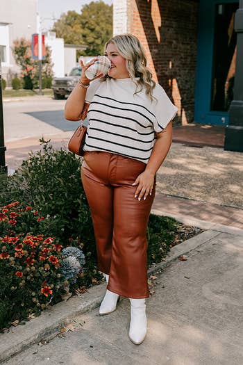 Model in striped top, brown leather pants, and white boots drinks from a cup while standing outside, holding a brown bag. Shopping article context