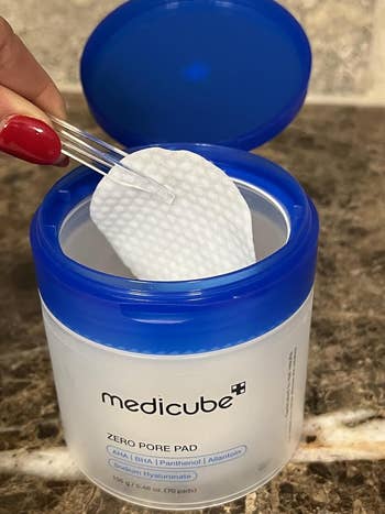 Hand utilizing tweezers to prime   a medicube Zero Pore Pad from an unfastened  container. The merchandise  is designed for skincare, featuring exfoliating ingredients