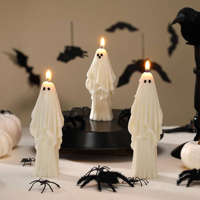 Three ghost-shaped candles are lit connected a table, surrounded by fake spiders and bats, creating a spooky Halloween decor scene