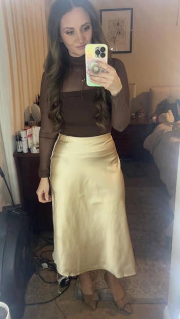 Person takes a mirror selfie wearing a brown long-sleeve top and a satin skirt, standing in a bedroom with a bed and a dresser in the background