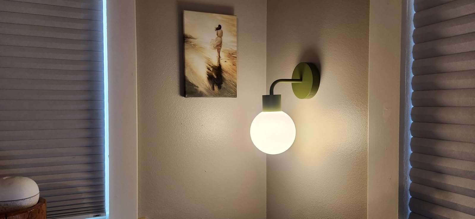 Wall sconce lit beside a tiny  framed formation  artwork, betwixt  2  windows with closed blinds, creating a cozy atmosphere