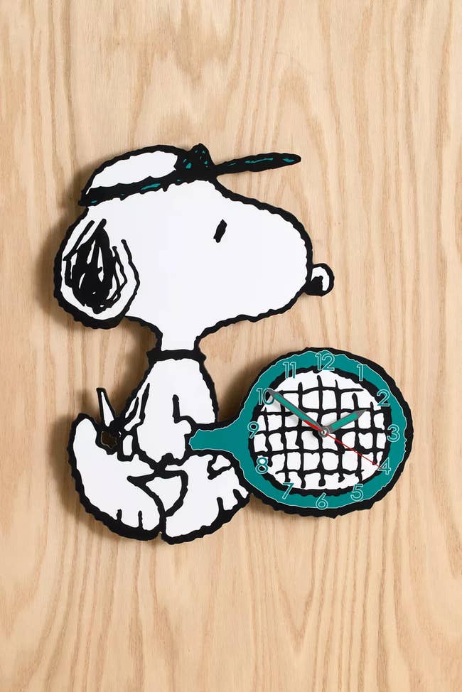 Wall timepiece featuring Snoopy holding a tennis racket, with timepiece hands connected the racket strings against a woody background