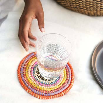 Hand placing a glass on a vibrant, beaded coaster on a tablecloth, next to dinnerware
