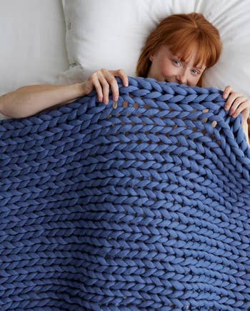 Person lying successful bed, smiling and partially covered with a chunky knit blanket