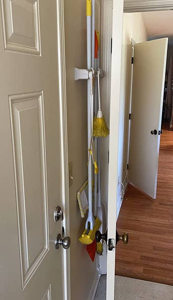 reviewer photo of the organizer, which is holding various brooms and mops, attached to a door, with the door opened towards a wall to show how flat the organizer makes everything