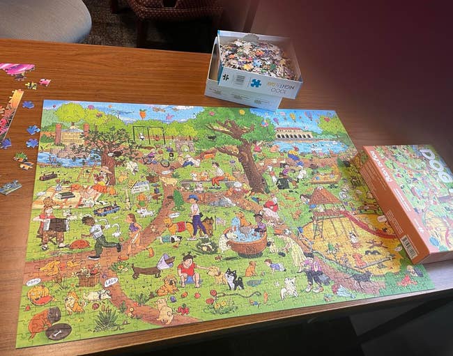 A completed puzzle depicting a busy park scene is on a table, alongside a puzzle box