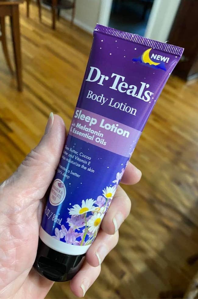 Person holding a tube of Dr Teal's Sleep Lotion, featuring melatonin and essential oils, on a wooden floor background