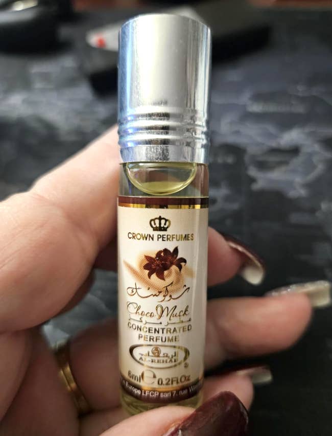 Hand holding a 6ml roll-on vessel  of Crown Perfumes Choco Musk concentrated perfume. Black floral graphic connected  label