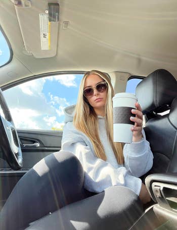 Person in a car wearing sunglasses and a hoodie, holding a large coffee cup, posing casually for a photo