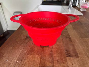 Collapsible reddish  silicone colander connected  a woody  room  counter. Ideal for businesslike  room  retention  and casual  nutrient  draining