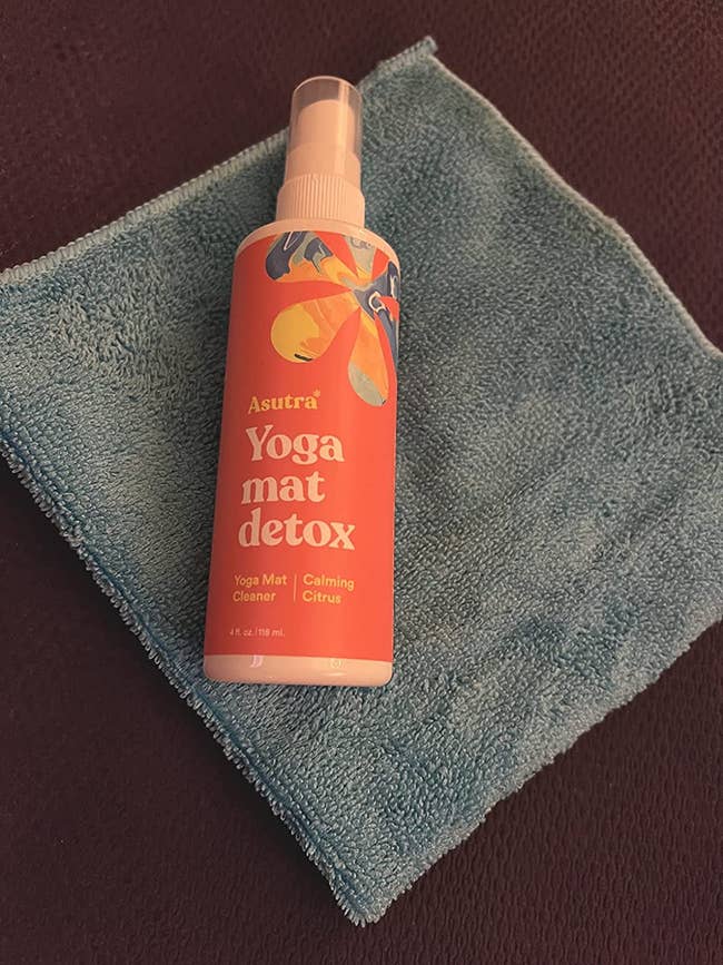 reviewer's spray bottle laying on a yoga mat with its cloth with an orange label with the words 