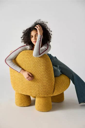 A person with curly hair sits thoughtfully on a textured, modern chair. They wear a long-sleeve top and wide-leg jeans