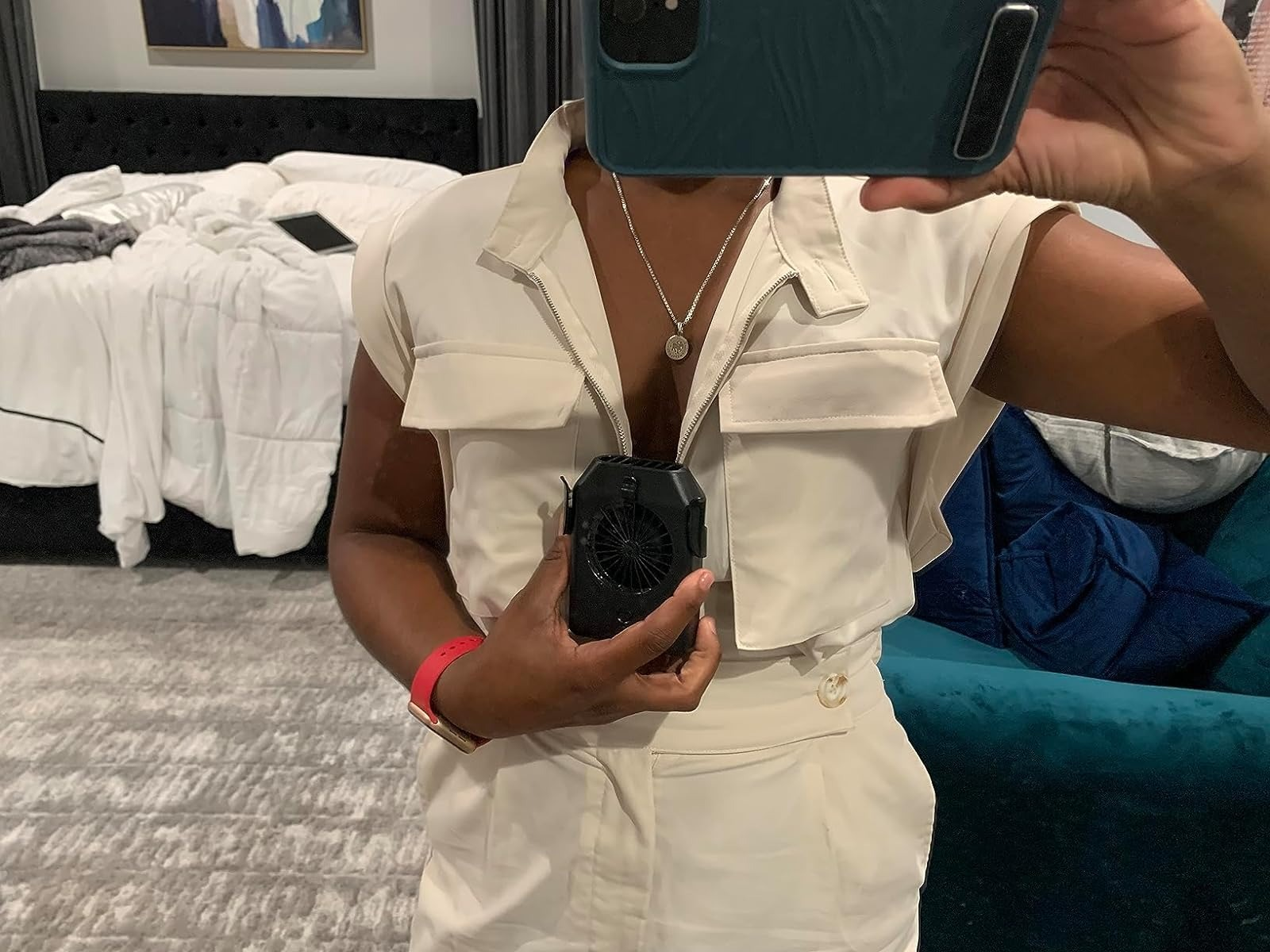 Orang yang mengambil selfie cermin di kamar tidur, mengenakan jumpsuit putih dan memegang kipas mini. Wajah tidak terlihat