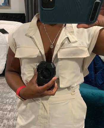Orang yang mengambil selfie cermin di kamar tidur, mengenakan jumpsuit putih dan memegang kipas mini. Wajah tidak terlihat