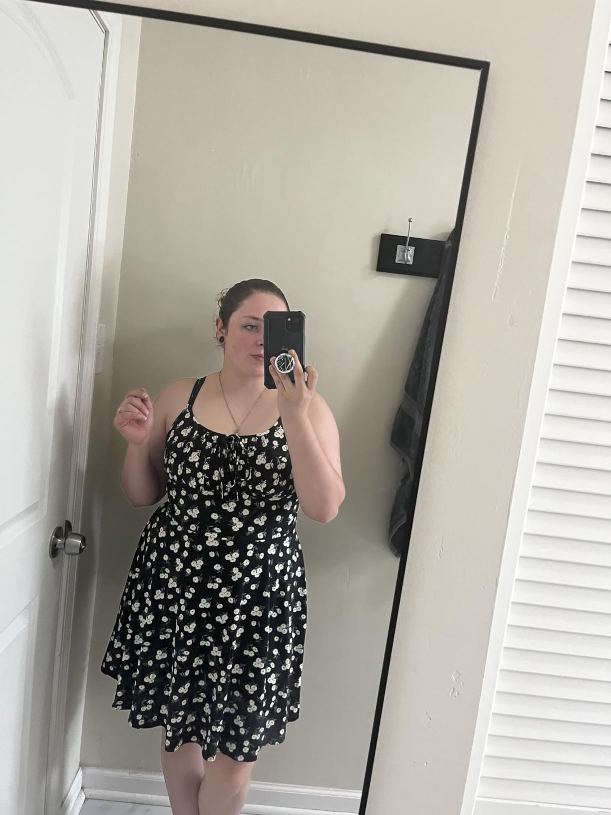 Person taking a reflector  selfie wearing a sleeveless floral dress, holding a phone. Room has a doorway  and a hanging hook successful  the background