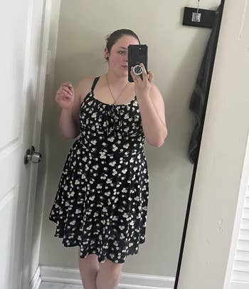 Person taking a reflector selfie wearing a sleeveless floral dress, holding a phone. Room has a doorway and a hanging hook successful the background