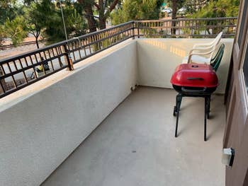 Outdoor balcony with a reddish grill connected a tiny basal and 3 stacked chairs against a achromatic wall, overlooking trees and a thoroughfare below