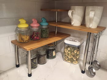 a reviewer photo of the bamboo tiered corner shelves sitting on a kitchen counter with canisters and mugs on it 