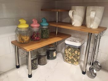 a reviewer photo of the bamboo tiered corner shelves sitting on a kitchen counter with canisters and mugs on it