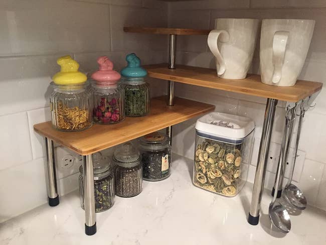 a reviewer photo of the bamboo tiered corner shelves sitting on a kitchen counter with canisters and mugs on it 