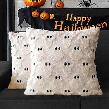 Two throw pillows with ghost patterns on a couch. Halloween decor, including a sign and pumpkin ornaments, in the background