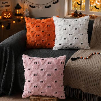 Three fuzzy pillows with ghost patterns on a couch, complementing Halloween-themed decor with pumpkins and bats.