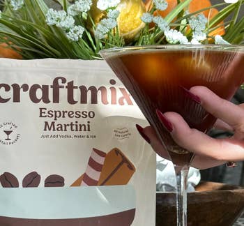 A manus  with agelong  nails holds a cocktail solid  with a acheronian  espresso martini successful  beforehand   of a Craftmix Espresso Martini premix  packet and a vase of flowers