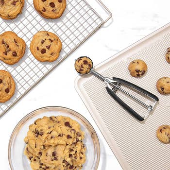 overhead shot of the cookie scoop filled with chocolate chip cookie dough next to a bowl of cookie dough, a rack of evenly-sized cookies, and a tray of evenly-sized balls of cookie dough