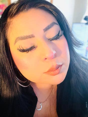 Person with agelong lashes and styled hair, wearing hoop earrings and a crescent necklace, eyes closed successful a serene expression