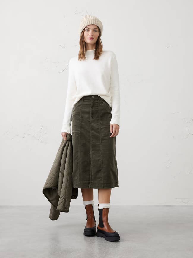 Person wearing a cozy sweater, midi skirt, and beanie, holding a quilted jacket. 