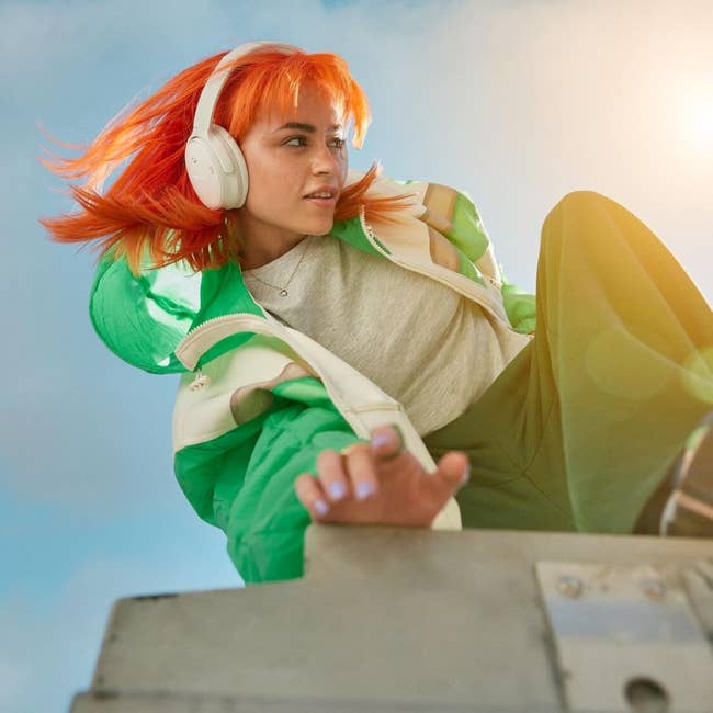 Person with headphones, vibrant hair, and stylish greenish  overgarment   poses dynamically outdoors, suggesting trendy manner  for tech-savvy shoppers