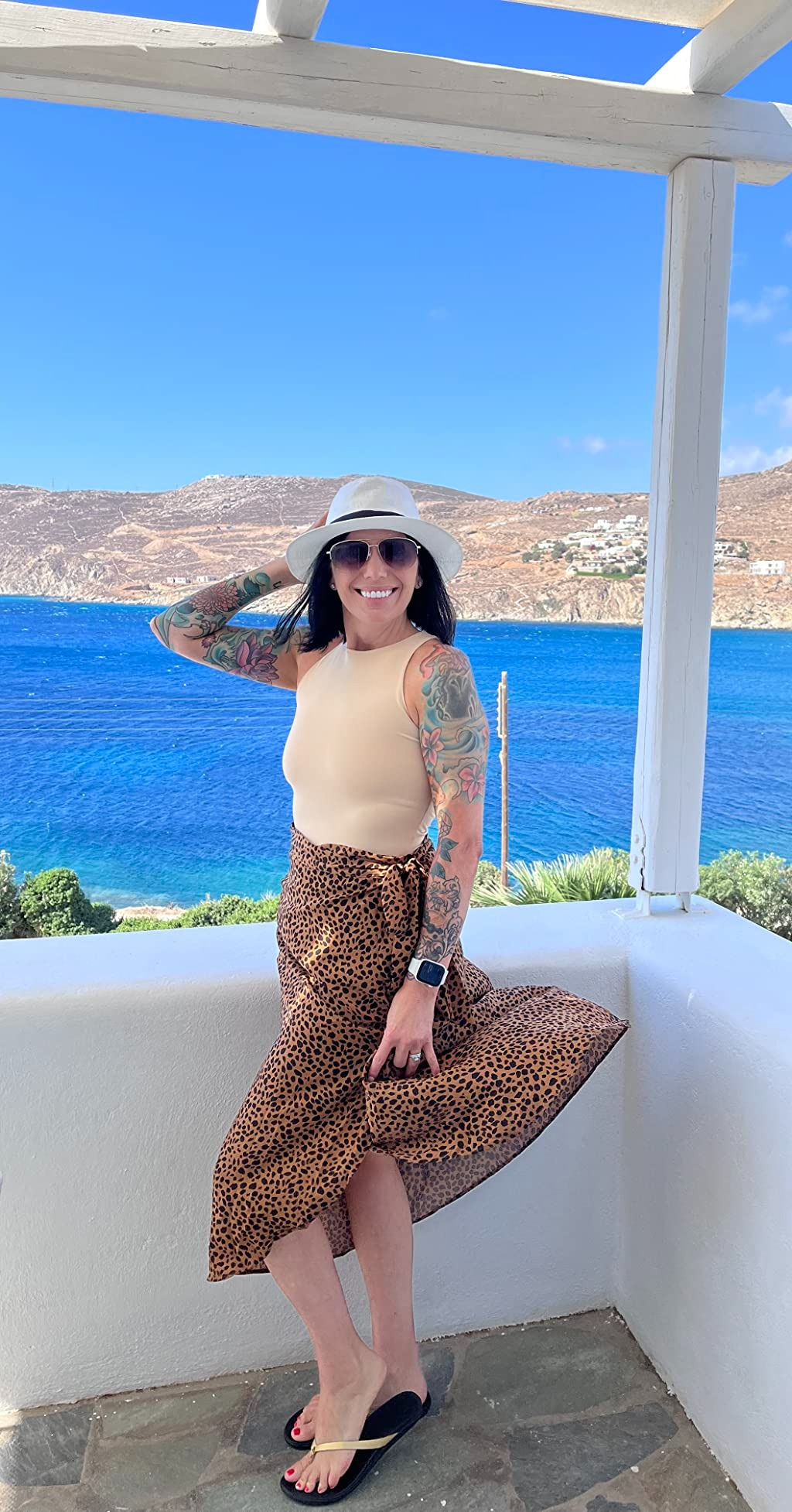 Wanita yang mengenakan atasan tanpa lengan, rok bermotif macan tutul, kacamata hitam, topi bertepi lebar, dan sandal berpose di teras dengan latar belakang pantai yang indah