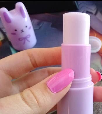 Close-up of a person's hand holding a twist-up lip balm with a cute bunny-shaped cap in the background. Their nails are neatly manicured